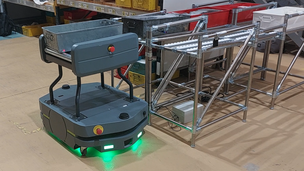 Autonomous vehicle alongside a conveyor transfer system in a manufacturing environment.